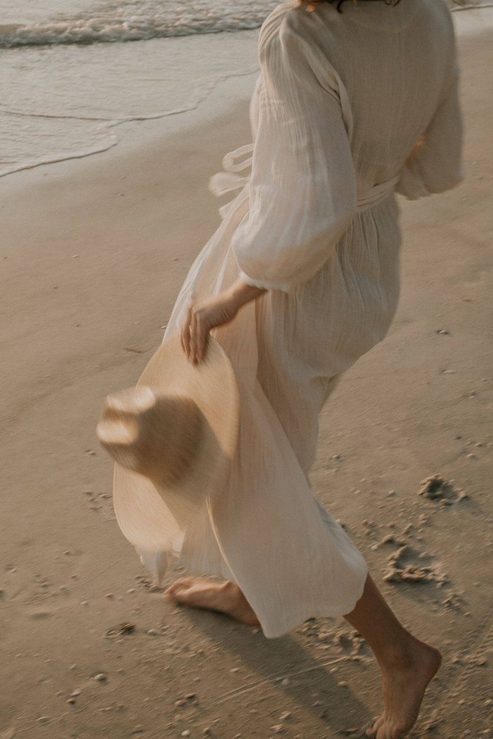 Person in a flowing white dress walking on a sandy beach.