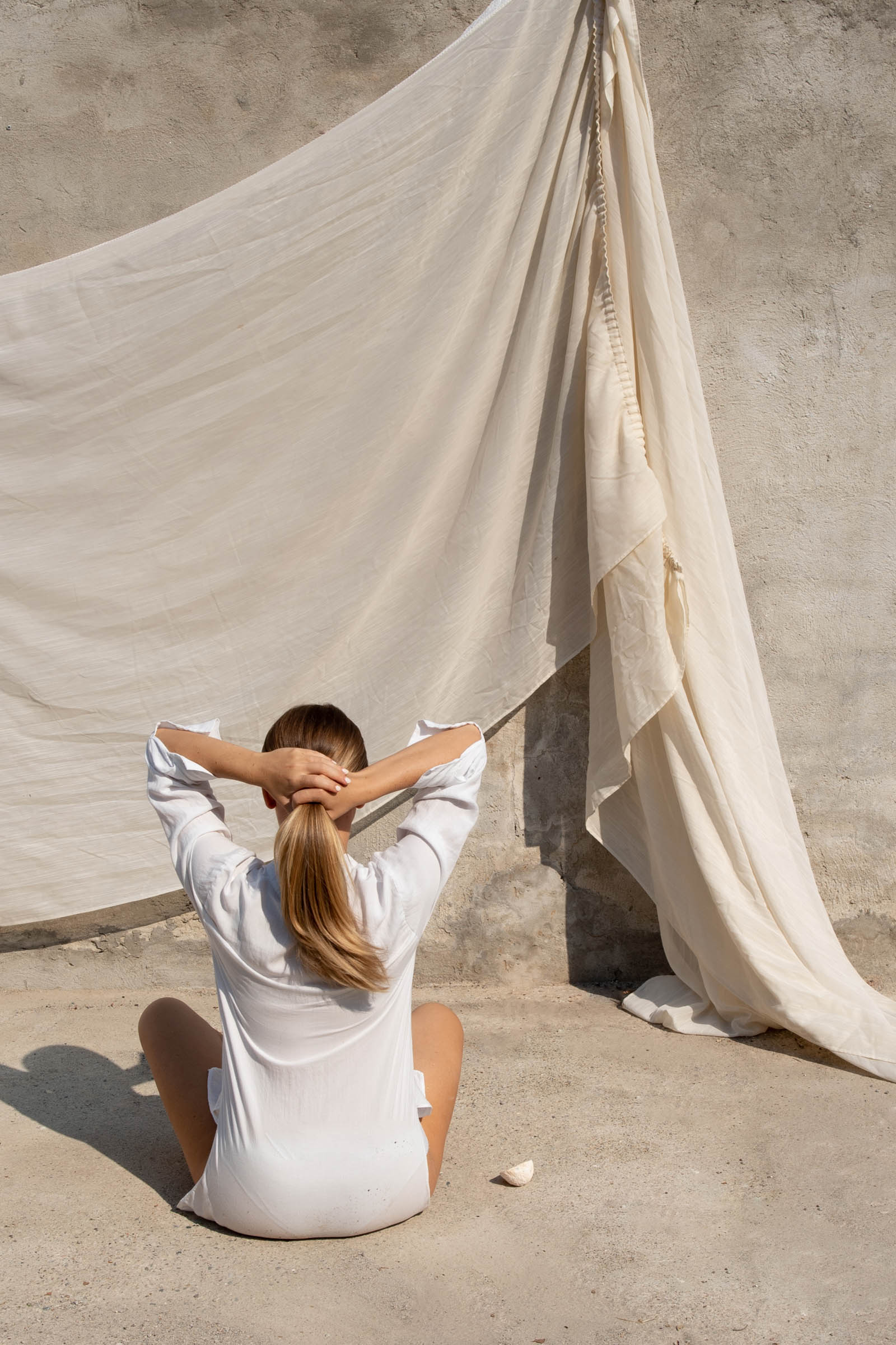 Woman with her back turned, gently holding her hair as linen curtains blow in the breeze—evoking calm, clarity, and a soft reset moment.